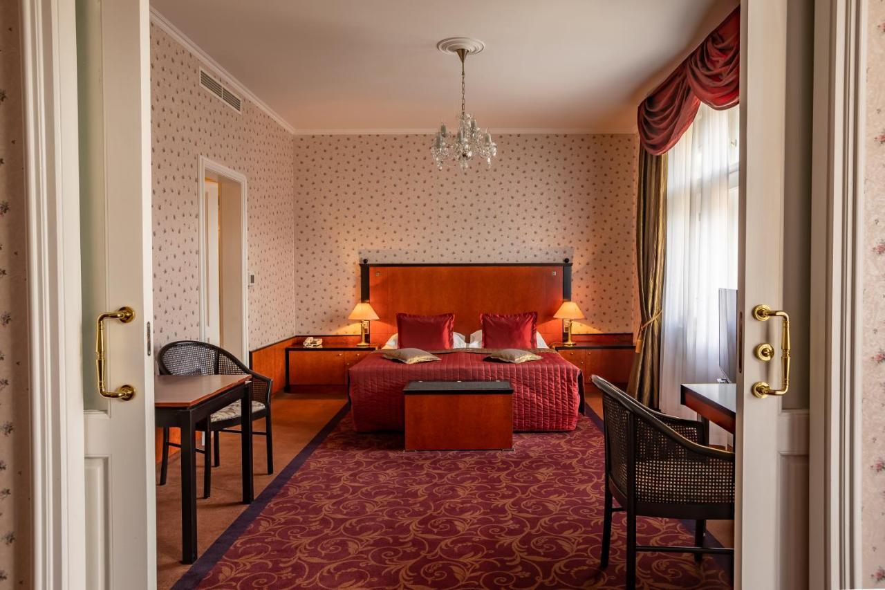 Grandhotel Pupp Κάρλοβι Βάρι Δωμάτιο φωτογραφία The photo shows the interior of a hotel room. Upon entering, you can see a spacious area with a plush, red carpet. The room features a large bed with decorative pillows and a wooden headboard, flanked by bedside tables with lamps. In front of the bed