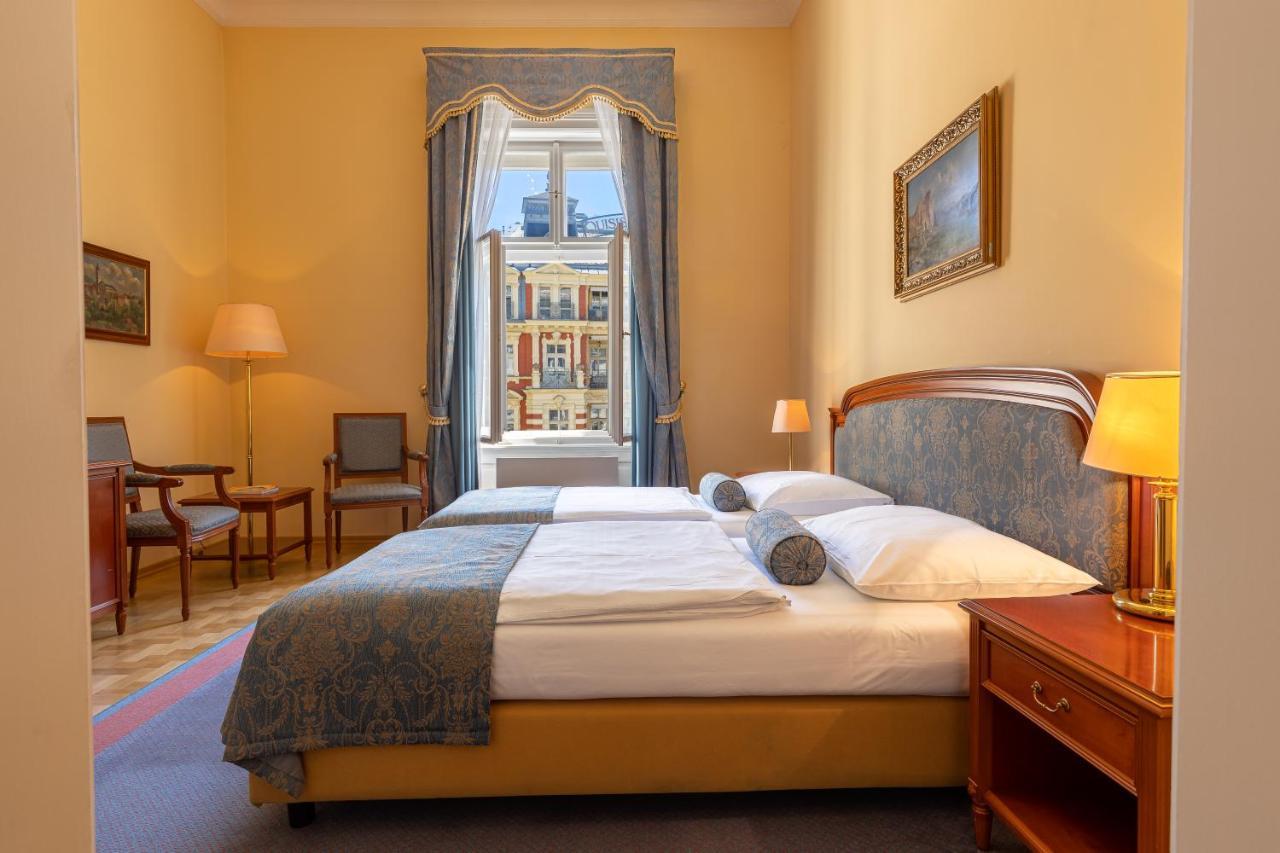 Grandhotel Pupp Κάρλοβι Βάρι Εξωτερικό φωτογραφία The photo depicts a cozy hotel room or bedroom. It features a neatly made bed with a blue and white color scheme, including decorative pillows. On one side of the bed is a small bedside table with a lamp. The room has warm yellow walls and a large wi