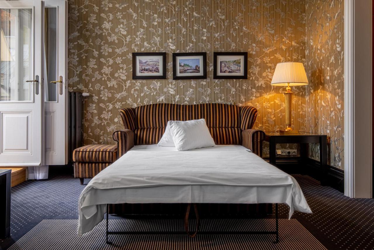 Grandhotel Pupp Κάρλοβι Βάρι Εξωτερικό φωτογραφία The photo depicts a cozy room featuring a striped sofa with a white pillow on it. In front of the sofa, there is a bed or a sleeping surface with a white sheet covering it. The walls are adorned with floral wallpaper, and there are three framed pictu