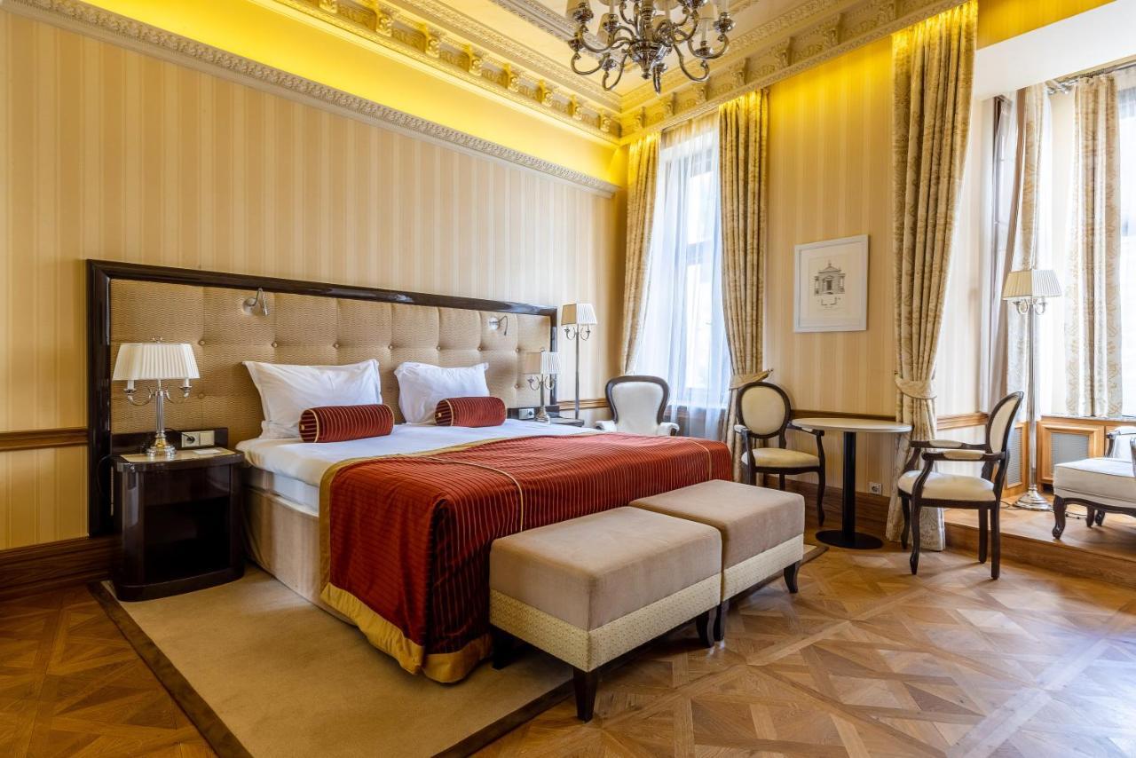 Grandhotel Pupp Κάρλοβι Βάρι Εξωτερικό φωτογραφία The photo shows a luxurious hotel room designed with an elegant interior. The room features a large bed with a tufted headboard, adorned with white bedding and a deep red throw. On either side of the bed, there are bedside lamps and a small seating a