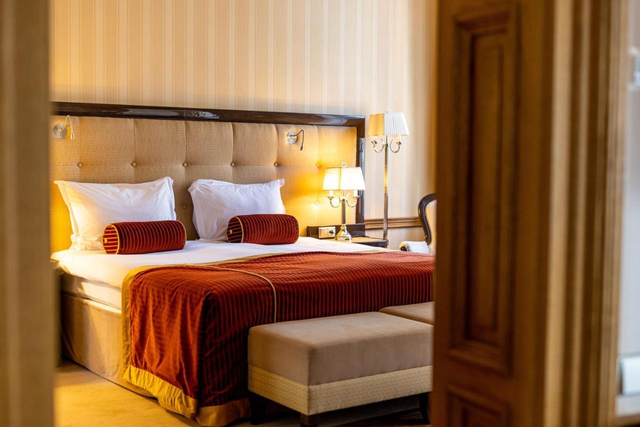 Grandhotel Pupp Κάρλοβι Βάρι Εξωτερικό φωτογραφία The photo shows a well-furnished hotel room with a cozy and elegant design. It features a large bed with white bedding and decorative orange-red throw pillows at the headboard. The walls are light-colored with subtle stripes, adding a touch of sophis
