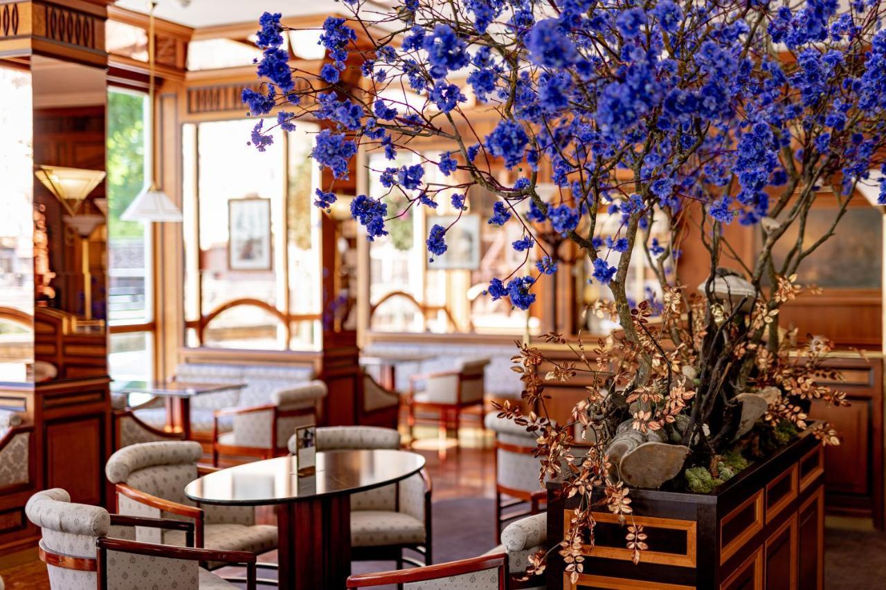 Grandhotel Pupp Κάρλοβι Βάρι Εξωτερικό φωτογραφία The photo depicts an elegant and stylish interior of a café or lounge. It features wooden furnishings with a warm color palette, including comfortable chairs and tables. A prominent feature is a large arrangement of blue flowers that adds a vibrant t