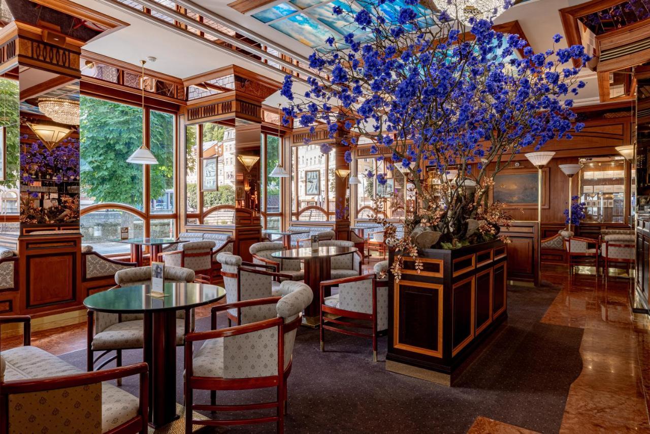 Grandhotel Pupp Κάρλοβι Βάρι Εξωτερικό φωτογραφία The photo shows an elegant interior of a restaurant or café. It features wooden furnishings with intricate details, including circular tables and upholstered chairs. A large floral arrangement, predominantly featuring blue flowers, is placed in the c