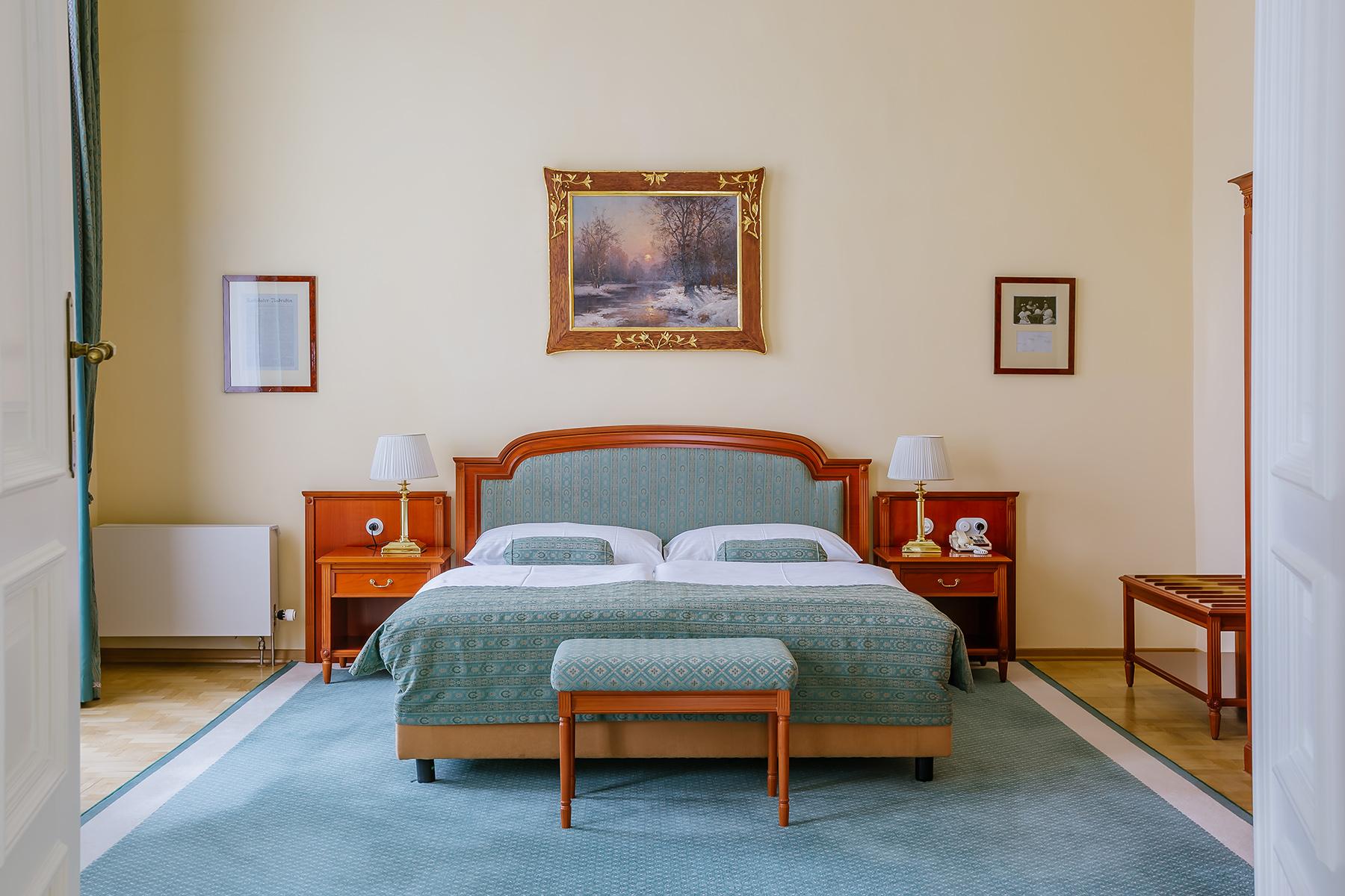 Grandhotel Pupp Κάρλοβι Βάρι Εξωτερικό φωτογραφία The photo shows a cozy bedroom with a light-colored wall. There is a large bed with a muted green coverlet and white bedding. On either side of the bed, there are bedside tables with lamps. At the foot of the bed, there is a small bench. Above the be