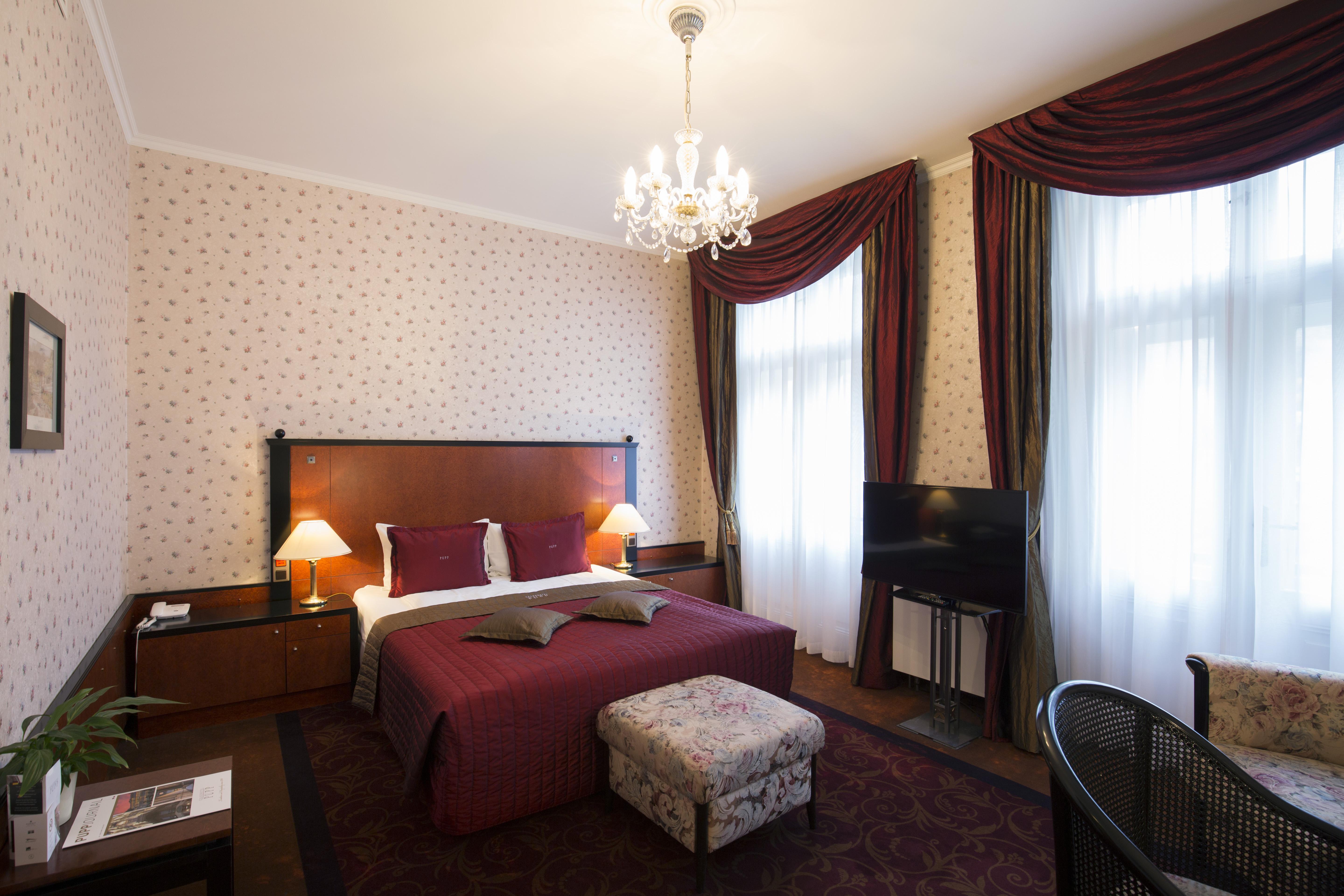 Grandhotel Pupp Κάρλοβι Βάρι Εξωτερικό φωτογραφία The photo shows a cozy hotel room with a double bed dressed in rich red bedding and two purple decorative pillows. The walls are adorned with a tasteful patterned wallpaper. There are two elegant windows with heavy, dark curtains that let in natural 
