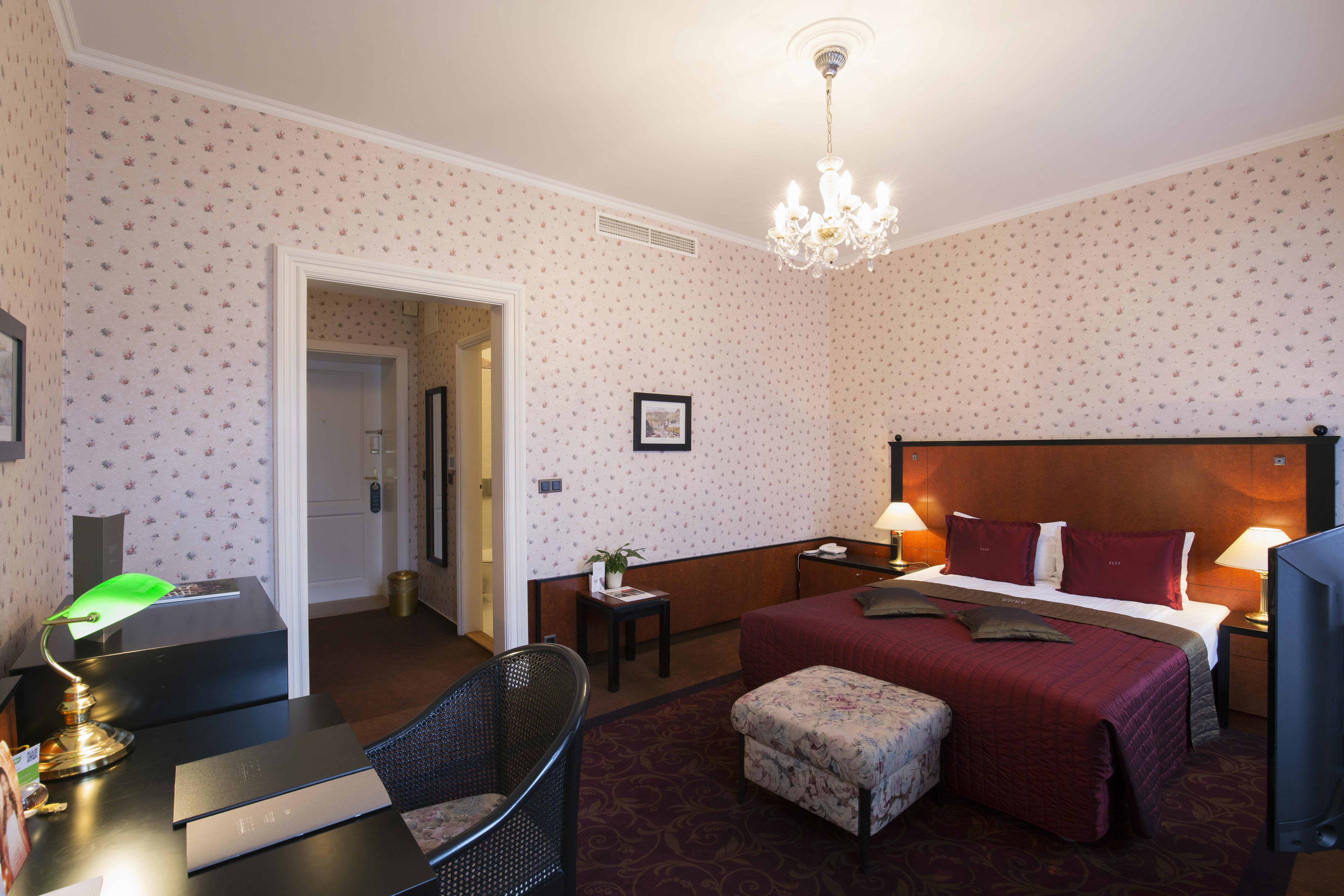 Grandhotel Pupp Κάρλοβι Βάρι Εξωτερικό φωτογραφία The photo depicts a well-furnished hotel room. There is a large bed with a burgundy bedspread, complemented by decorative pillows. The room features soft lighting from a chandelier and lamps on either side of the bed. 

To the left, there is a desk w