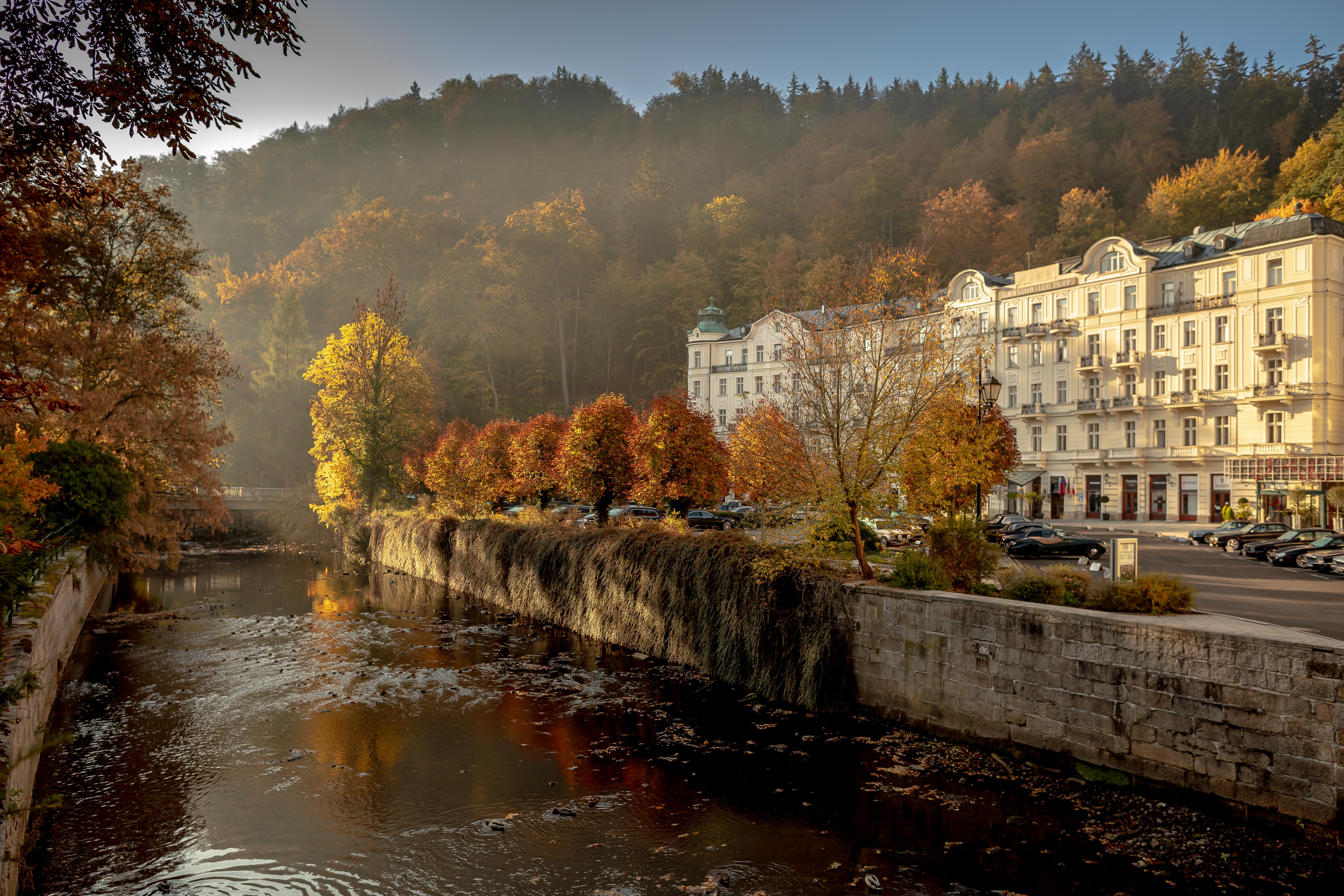 Grandhotel Pupp Κάρλοβι Βάρι Εξωτερικό φωτογραφία The photo depicts a serene riverside scene during autumn. The river flows gently, reflecting the warm hues of the trees along its banks. On one side, there are vibrant orange and yellow trees, showcasing the beautiful fall foliage, while the opposite