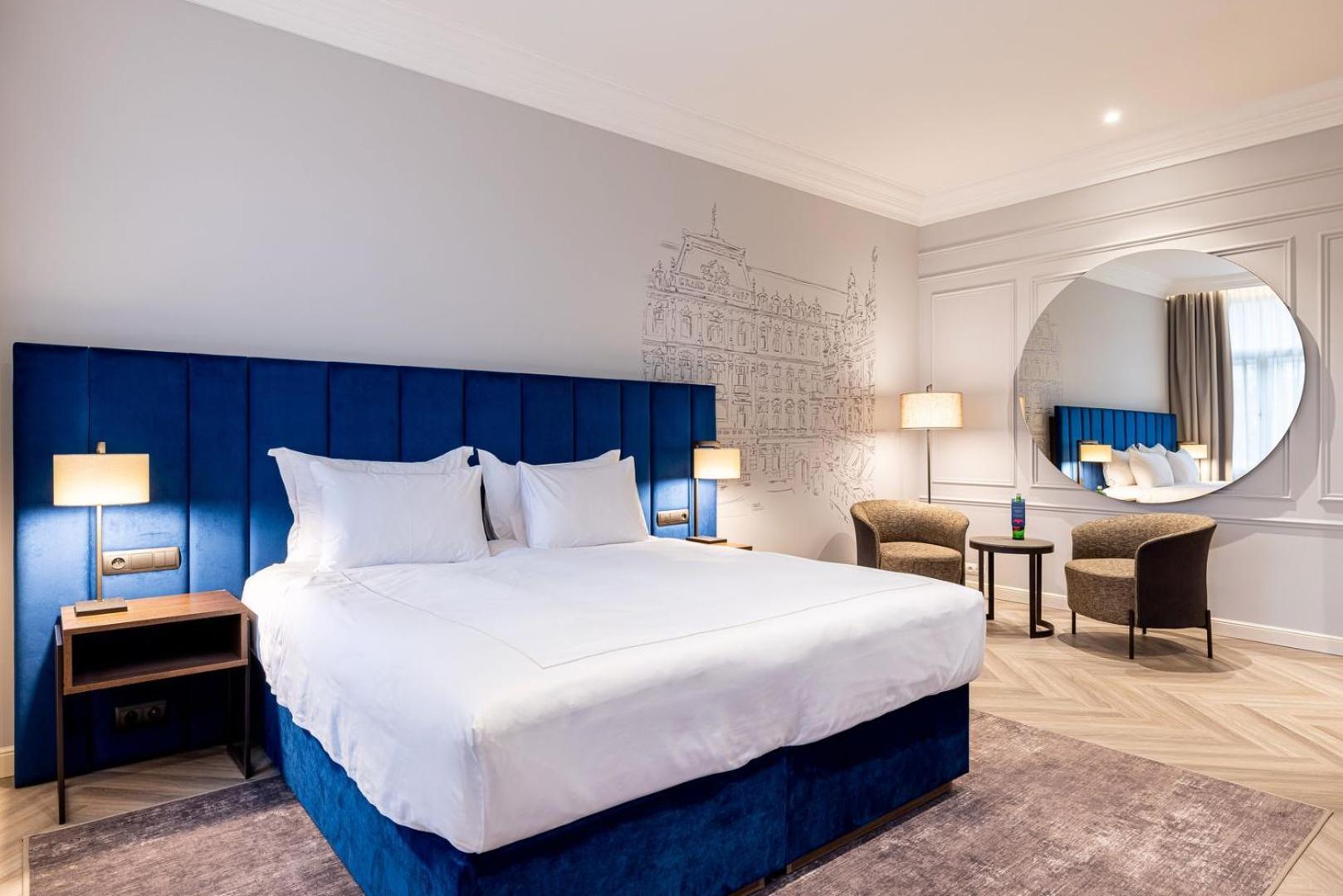 Grandhotel Pupp Κάρλοβι Βάρι Εξωτερικό φωτογραφία The photo shows a modern hotel room featuring a large double bed with white linens and a blue upholstered headboard. The walls are light gray, and there is a decorative mural in the background. Two bedside lamps are on either side of the bed. In the 