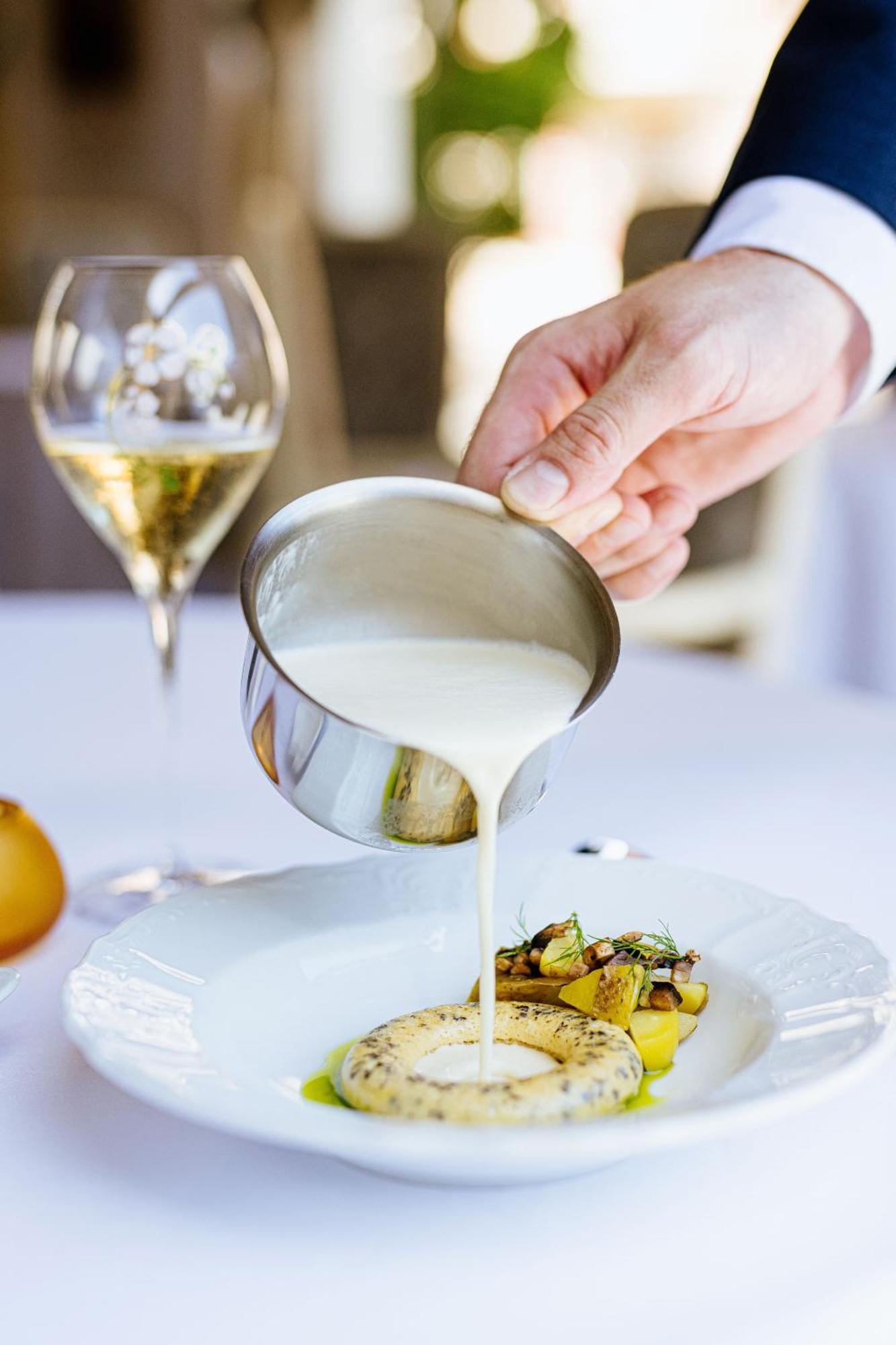 Grandhotel Pupp Κάρλοβι Βάρι Εξωτερικό φωτογραφία The photo shows a hand pouring a creamy sauce from a small stainless steel container over a plated dish, which appears to include vegetables and possibly pasta. In the background, there's a glass of sparkling wine, suggesting a fine dining setting. T