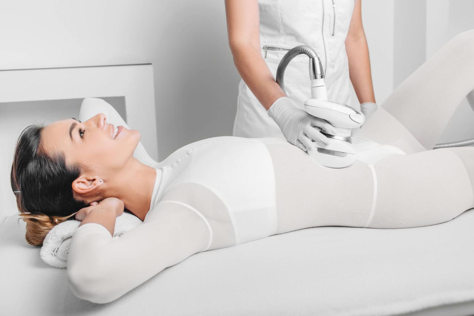Grandhotel Pupp Κάρλοβι Βάρι Εξωτερικό φωτογραφία The photo depicts a woman lying on a treatment table, appearing relaxed and smiling. She is wearing a form-fitting white outfit, likely designed for a cosmetic or wellness procedure. A technician, dressed in a white outfit and gloves, is applying a d