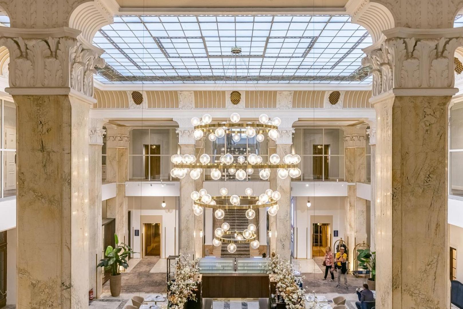 Grandhotel Pupp Κάρλοβι Βάρι Εξωτερικό φωτογραφία The photo depicts an elegant hotel lobby characterized by its spacious and luxurious design. Large marble columns rise to support a ceiling with a glass skylight, allowing natural light to illuminate the area. A central chandelier made of numerous ro